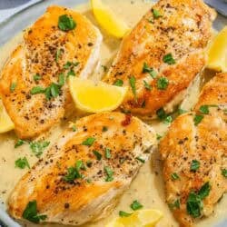 Four garlic parmesan chicken fillets topped with sauce and parsley on a plate with lemon wedges.