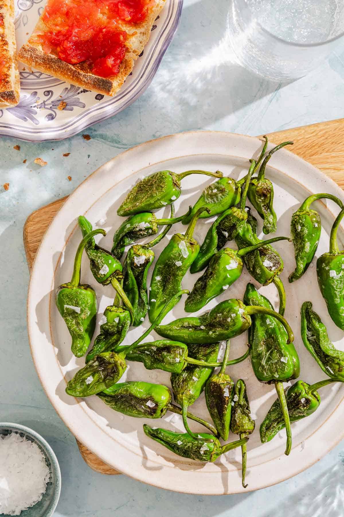 Padron peppers topped with flakey sea salt on a plate. Next to this is a a glass of water, a bowl of the salt and a plate of pan con tomate.
