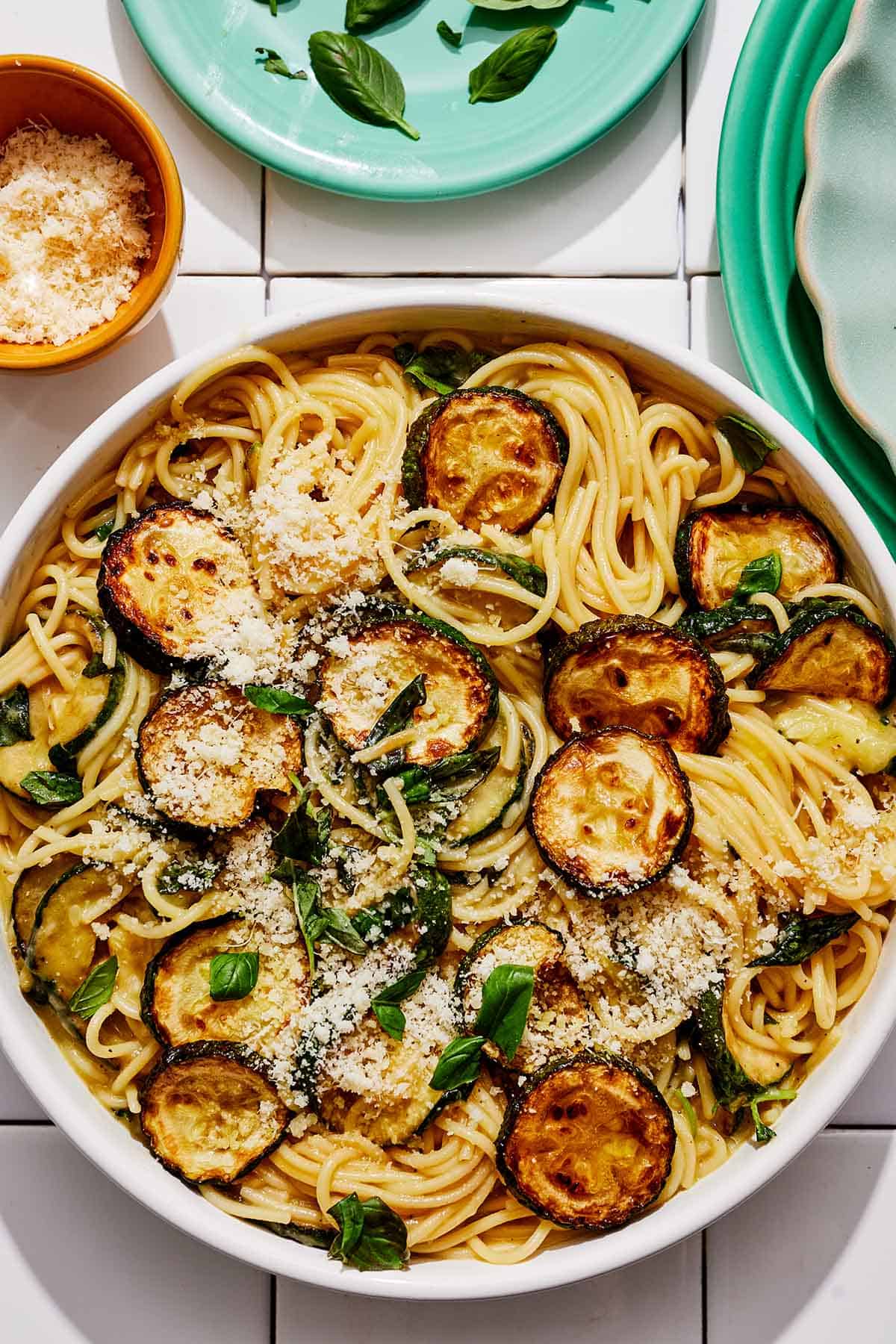 Spaghetti alla nerano in a serving bowl next to a plate of basil, and a bowl of grated parmagiano reggiano cheese.