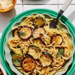 A serving of spaghetti alla nerano on a plate next to a plate of basil and a bowl of grated Parmagiano Reggiano cheese.