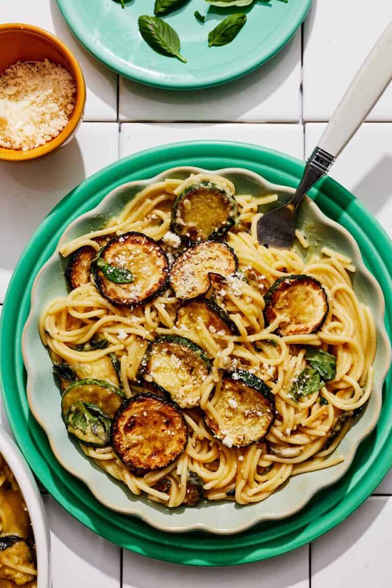 A serving of spaghetti alla nerano on a plate next to a plate of basil and a bowl of grated Parmagiano Reggiano cheese.