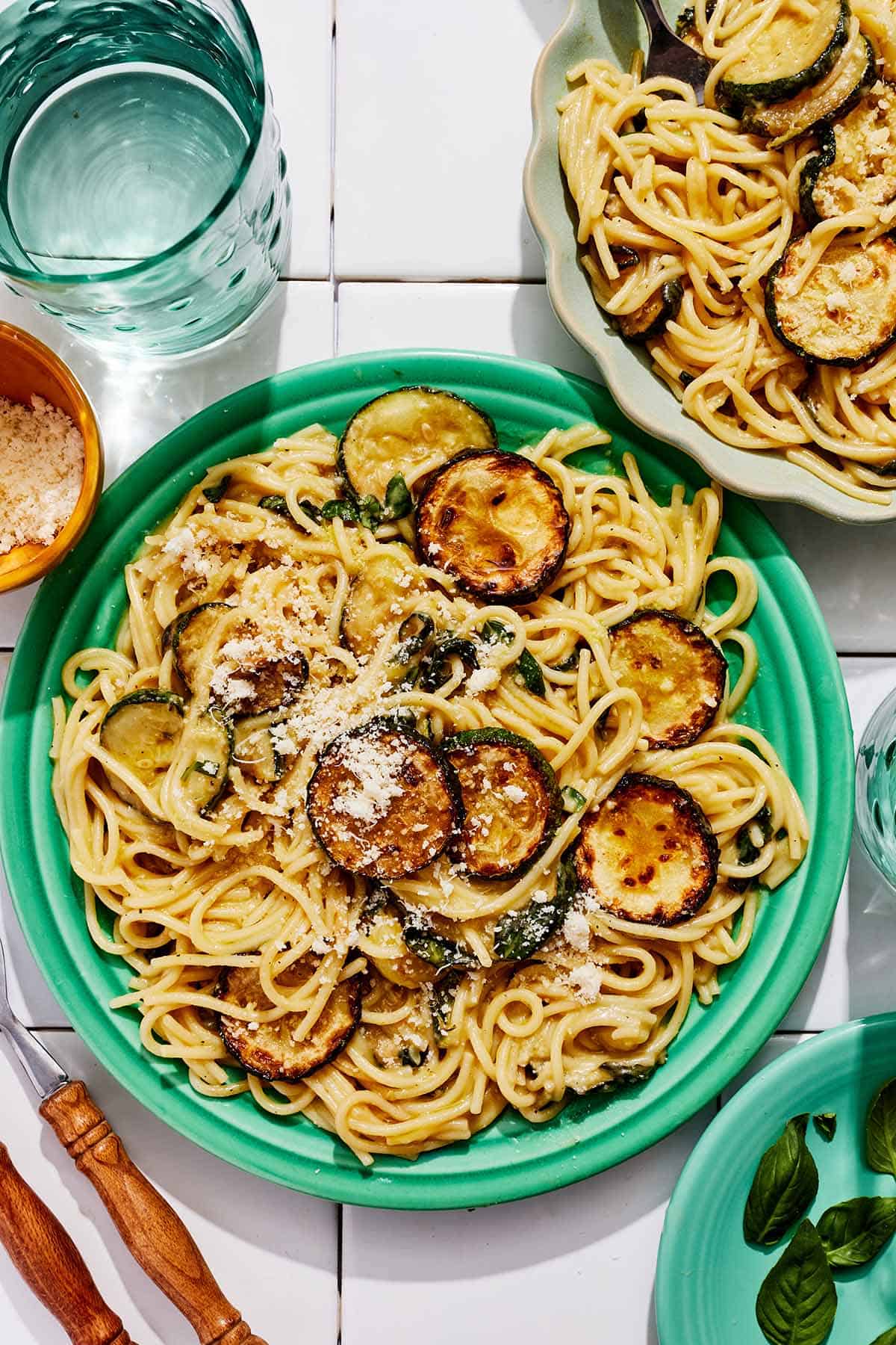 A serving of spaghetti alla nerano on a plate. Next to this is the rest of the recipe in a bowl, a glass of water, and a bowl of grated Parmagiano Reggiano cheese.