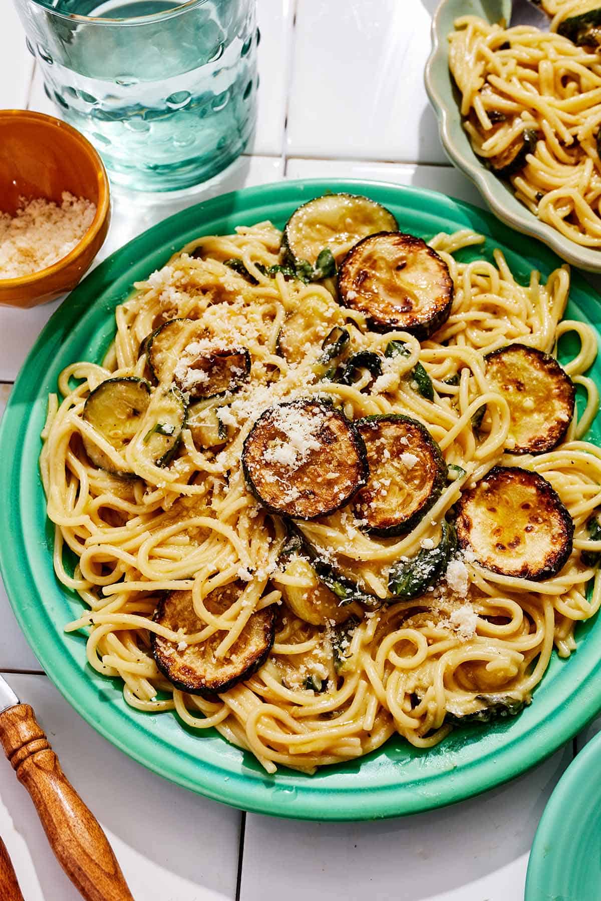 A serving of spaghetti alla nerano on a plate. Next to this is the rest of the recipe in a bowl, a glass of water, and a bowl of grated Parmagiano Reggiano cheese.