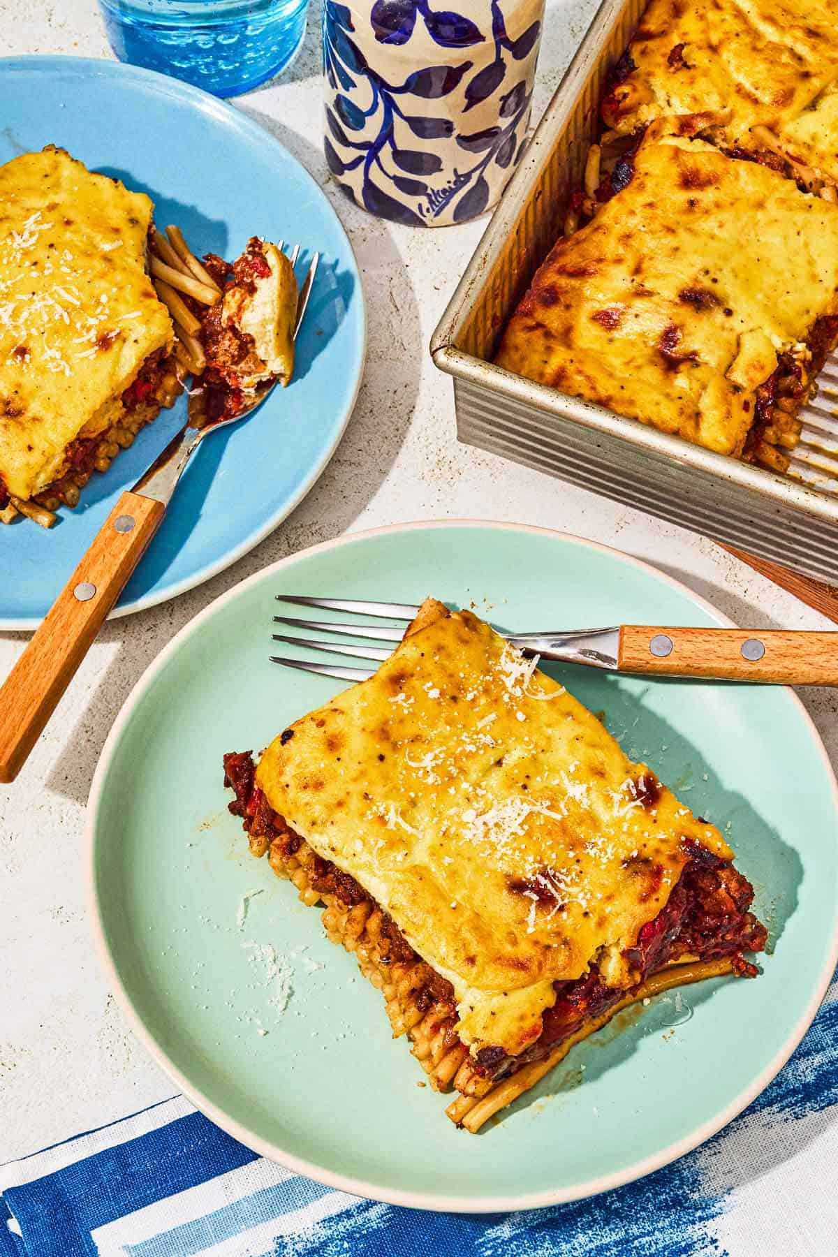 Two slices of pastitsio on plates with forks next to the rest of the pastitsio recipe in a baking dish.