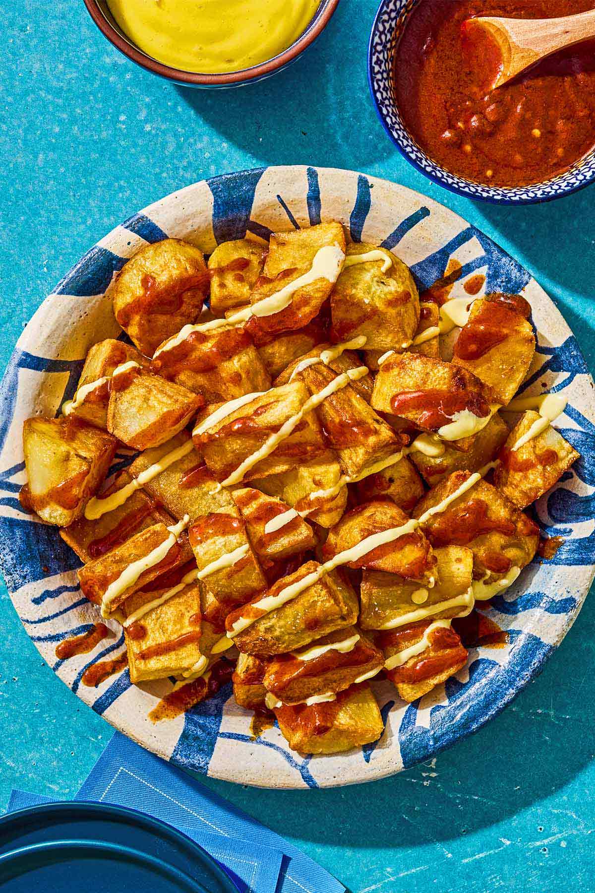 Fried potatoes for the patatas bravas in a serving bowl next to bowls of bravas sauce and aioli.