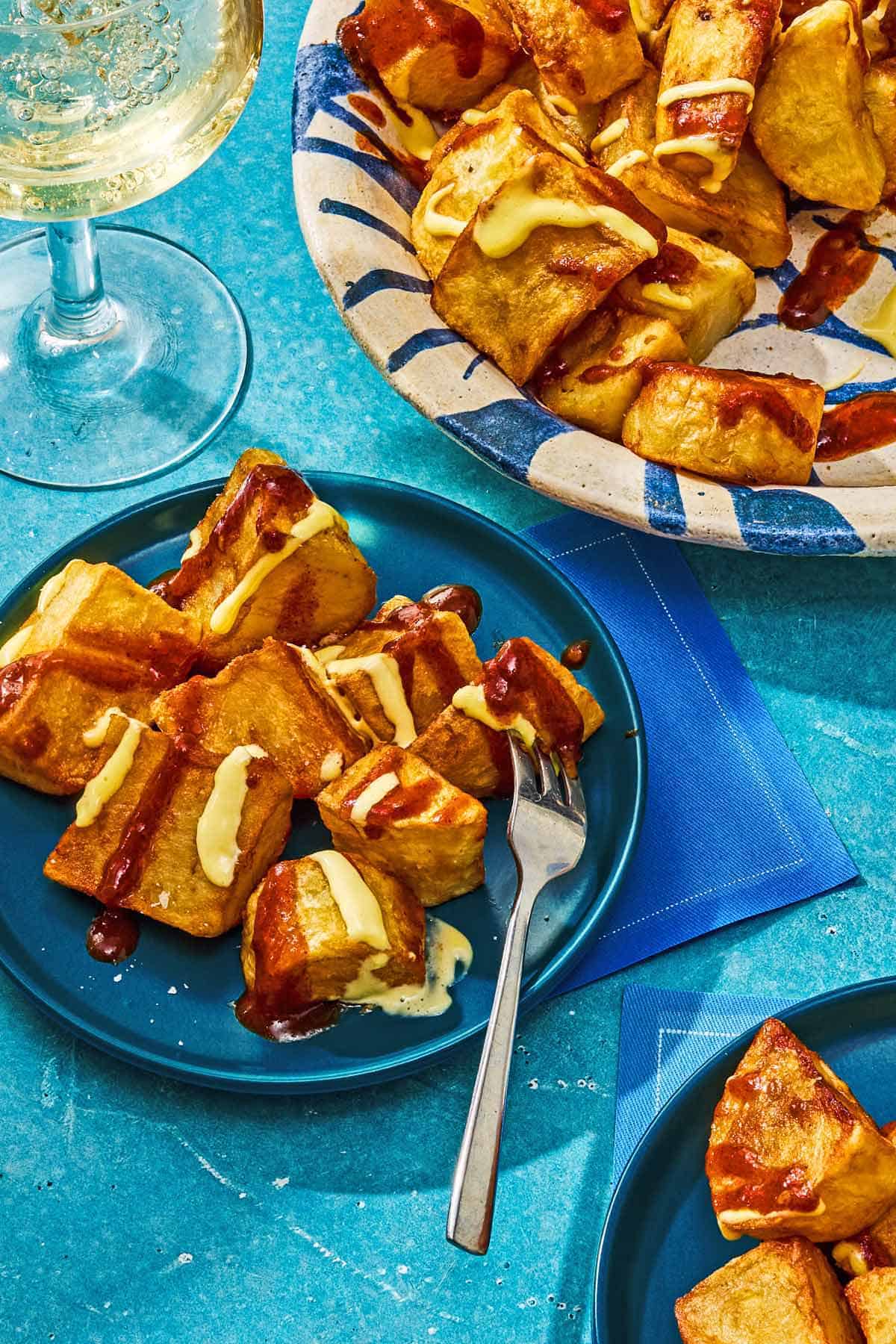 A serving of patatas bravas on a plate with a fork. Next to this is the rest of the recipe in a serving bowl and a glass of wine.