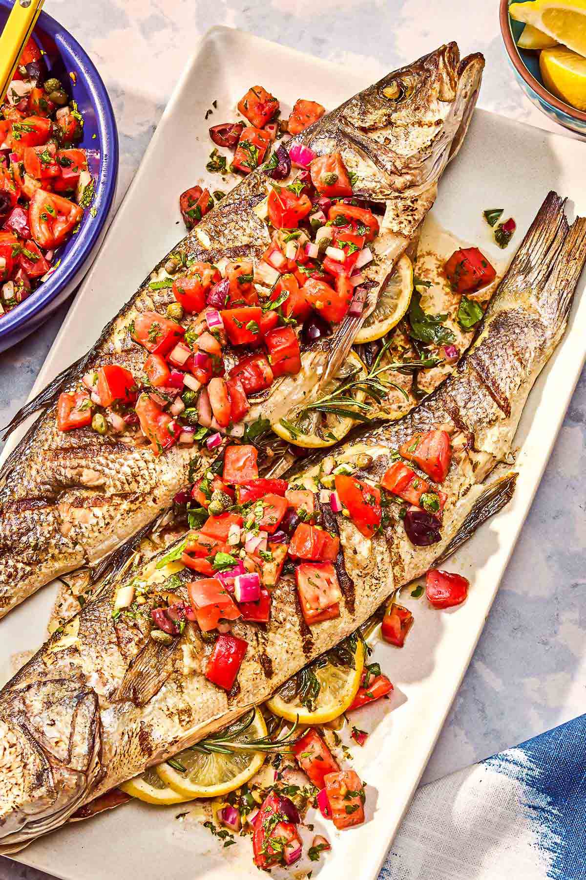 Two grilled branzino topped with tomato caper salsa on a platter next to a bowl of the salsa.