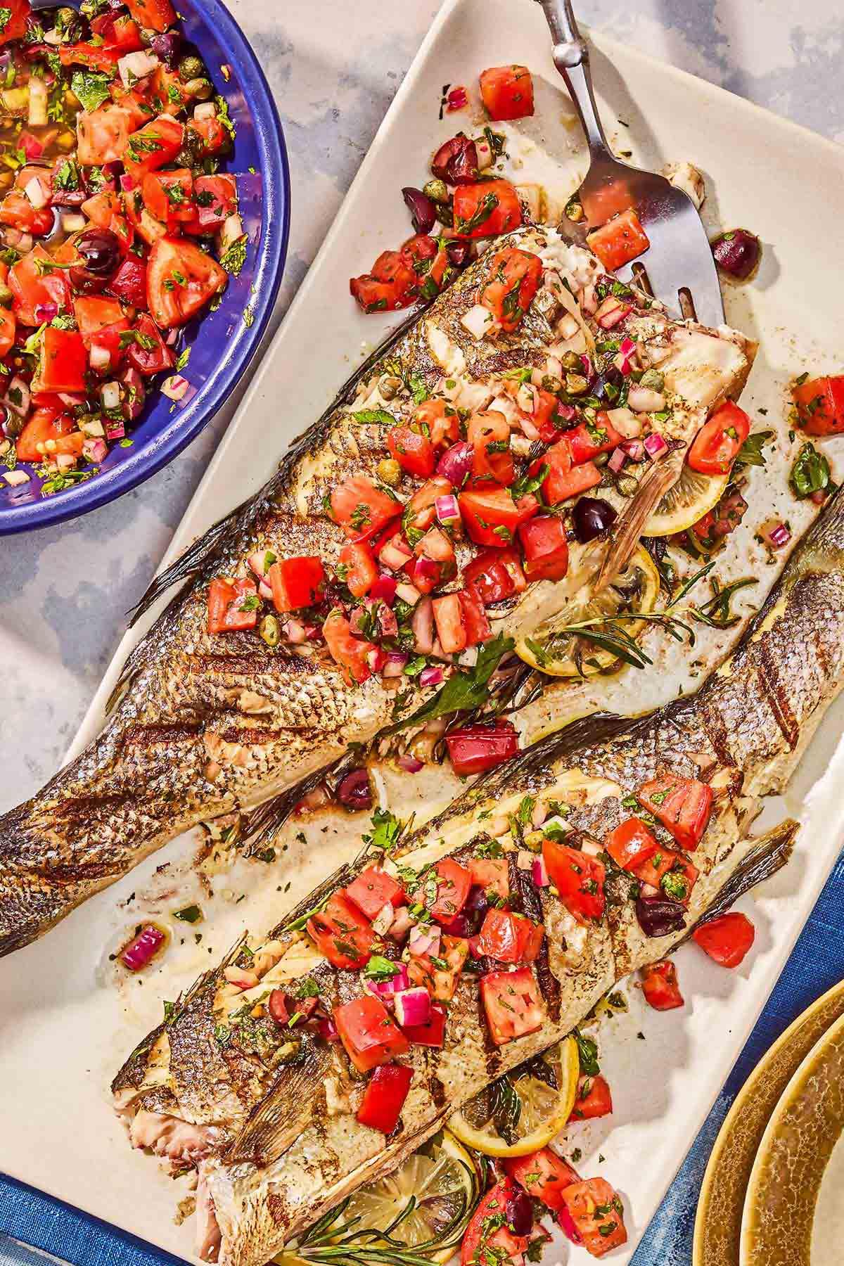 Two grilled branzino topped with tomato caper salsa on a platter next to a bowl of the salsa.