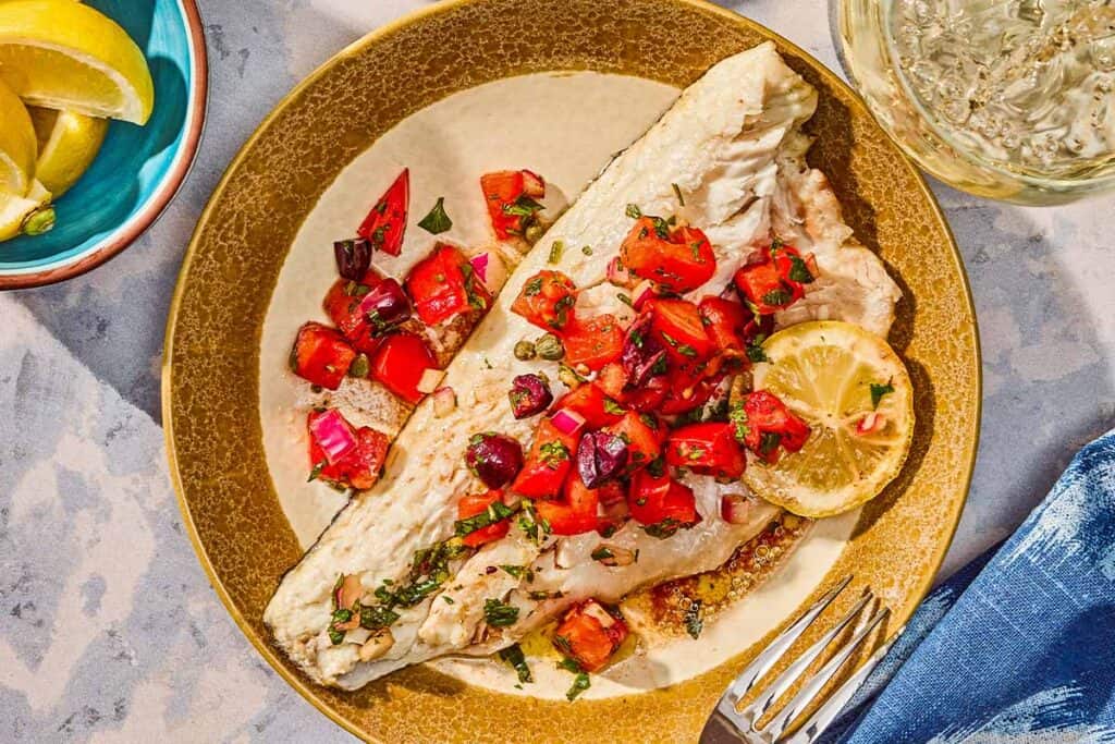 A grilled branzino fillet topped with tomato-caper sauce and a lemon wheel on a plate with a fork. Next to this is a bowl of lemon wedges and a glass of wine