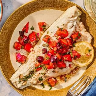 A grilled branzino fillet topped with tomato-caper sauce and a lemon wheel on a plate with a fork. Next to this is a bowl of lemon wedges and a glass of wine