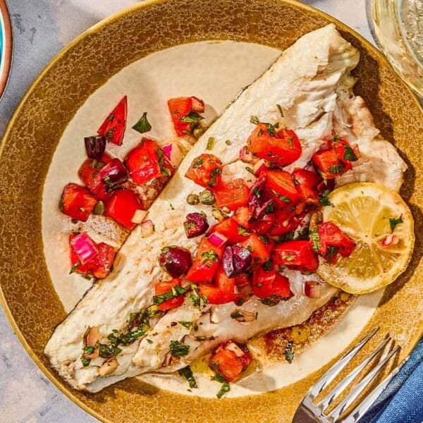 A grilled branzino fillet topped with tomato-caper sauce and a lemon wheel on a plate with a fork. Next to this is a bowl of lemon wedges and a glass of wine