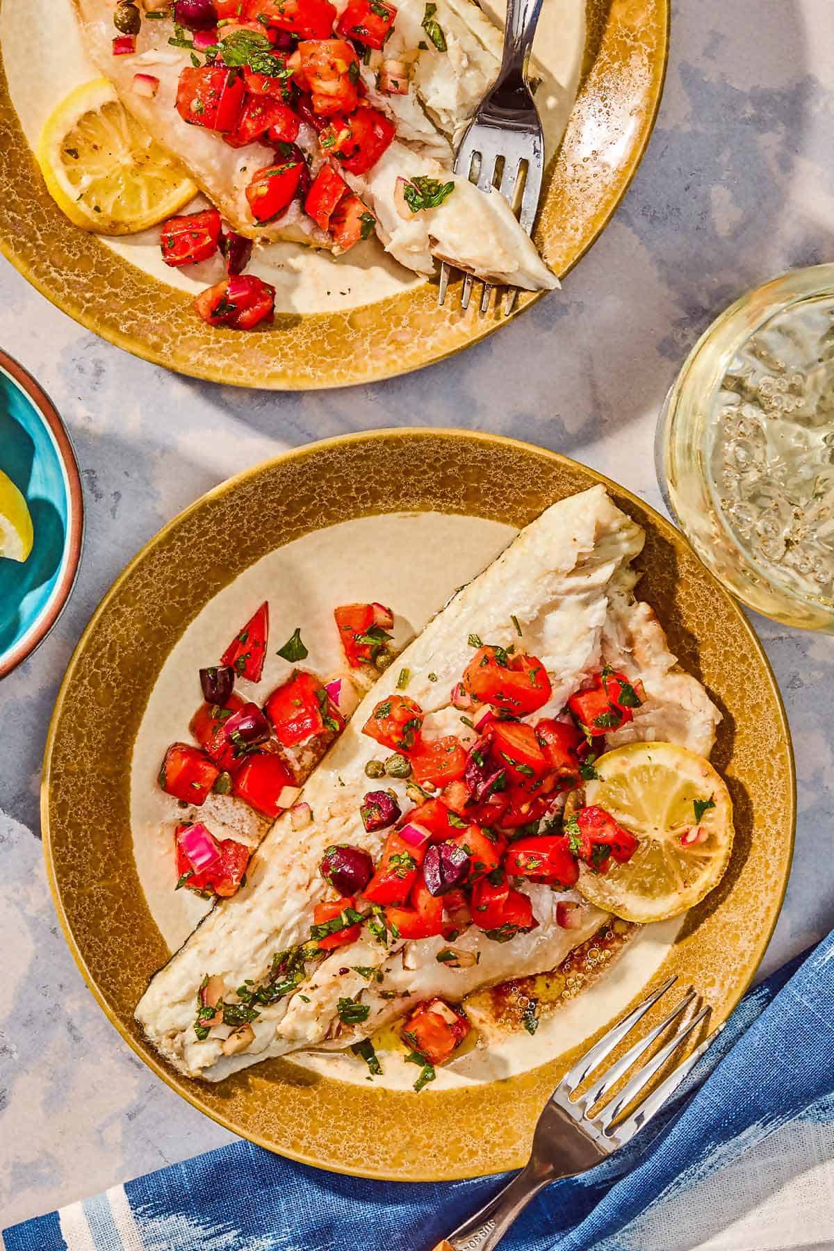 2 grilled branzino fillets topped with tomato-caper sauce and a lemon wheel on plates with forks next to a glass of wine.