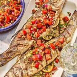 Two grilled branzino topped with tomato caper salsa on a platter. Next to this is a bowl of the salsa, a stack of 2 plates and a glass of wine.