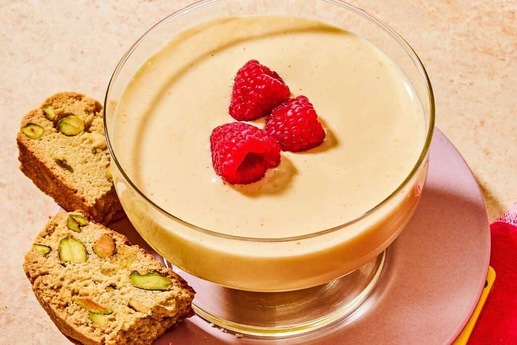 Zabaglione in a bowl topped with raspberries on a plate with biscotti.