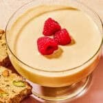 Zabaglione in a bowl topped with raspberries on a plate with biscotti.