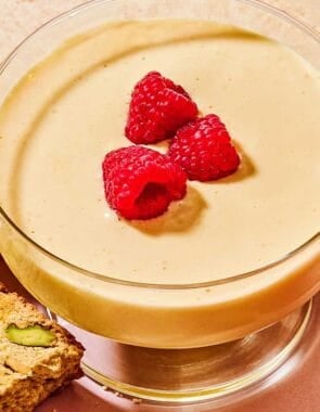 Zabaglione in a bowl topped with raspberries on a plate with biscotti.