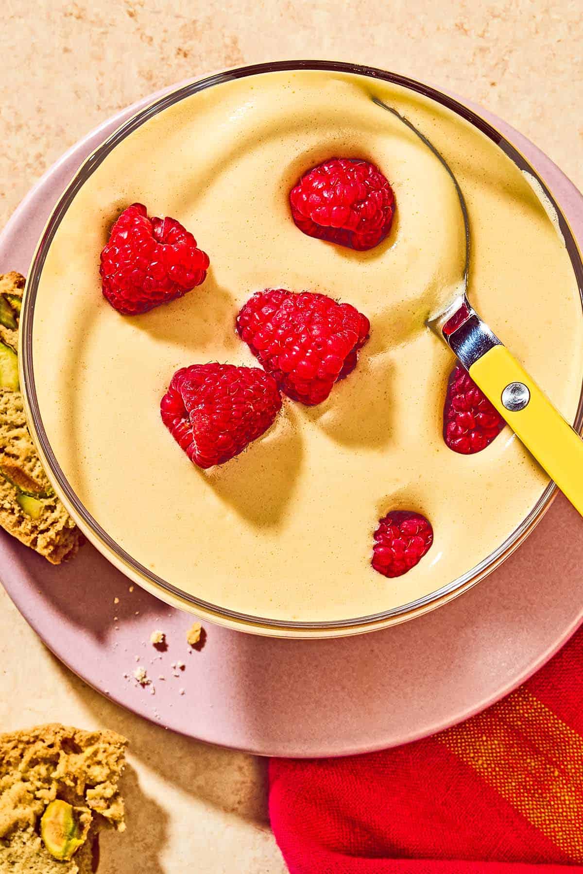 Zabaglione in a bowl topped with raspberries on a plate with biscotti.