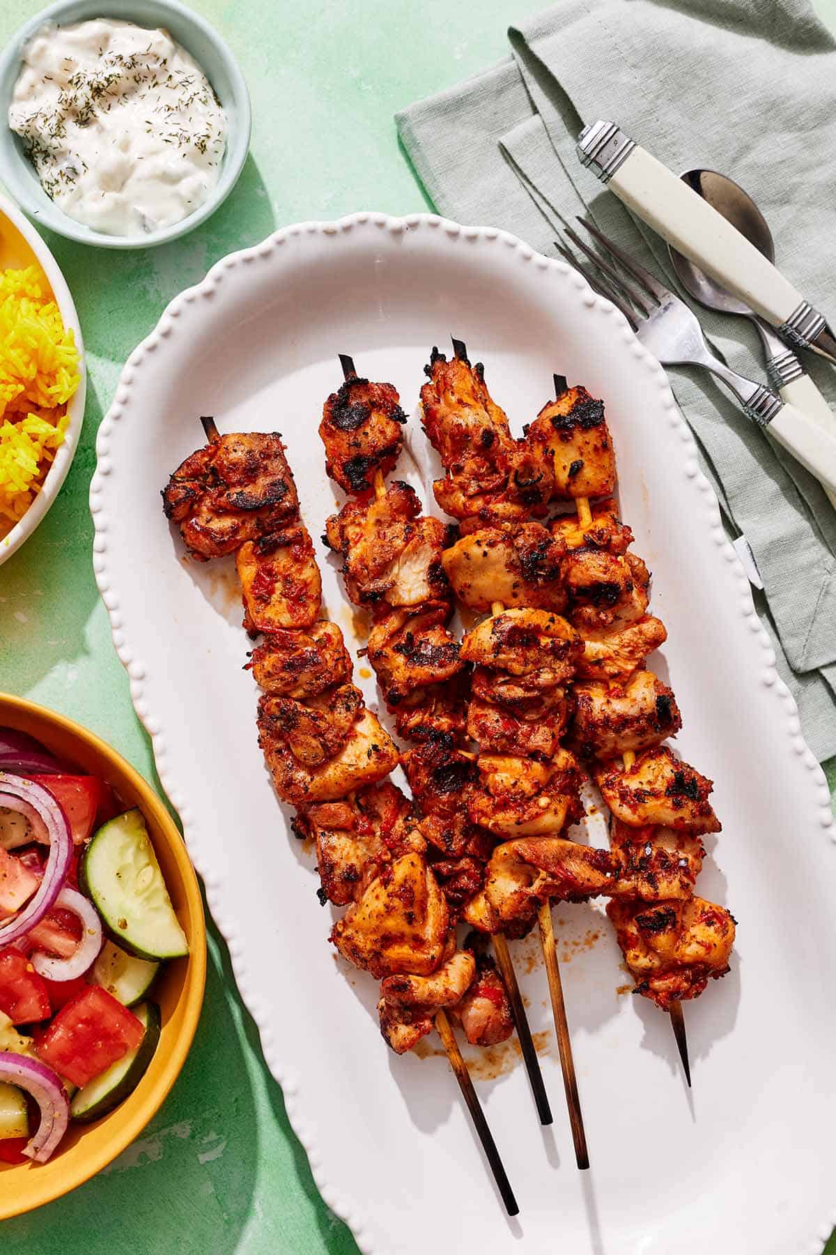 Turkish chicken skewers on a platter next to utensils, and bowls of tzatziki, rice and a cucumber tomato salad.