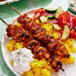 Turkish chicken skewers on a plate with rice, tzatziki and a cucumber and tomato salad.
