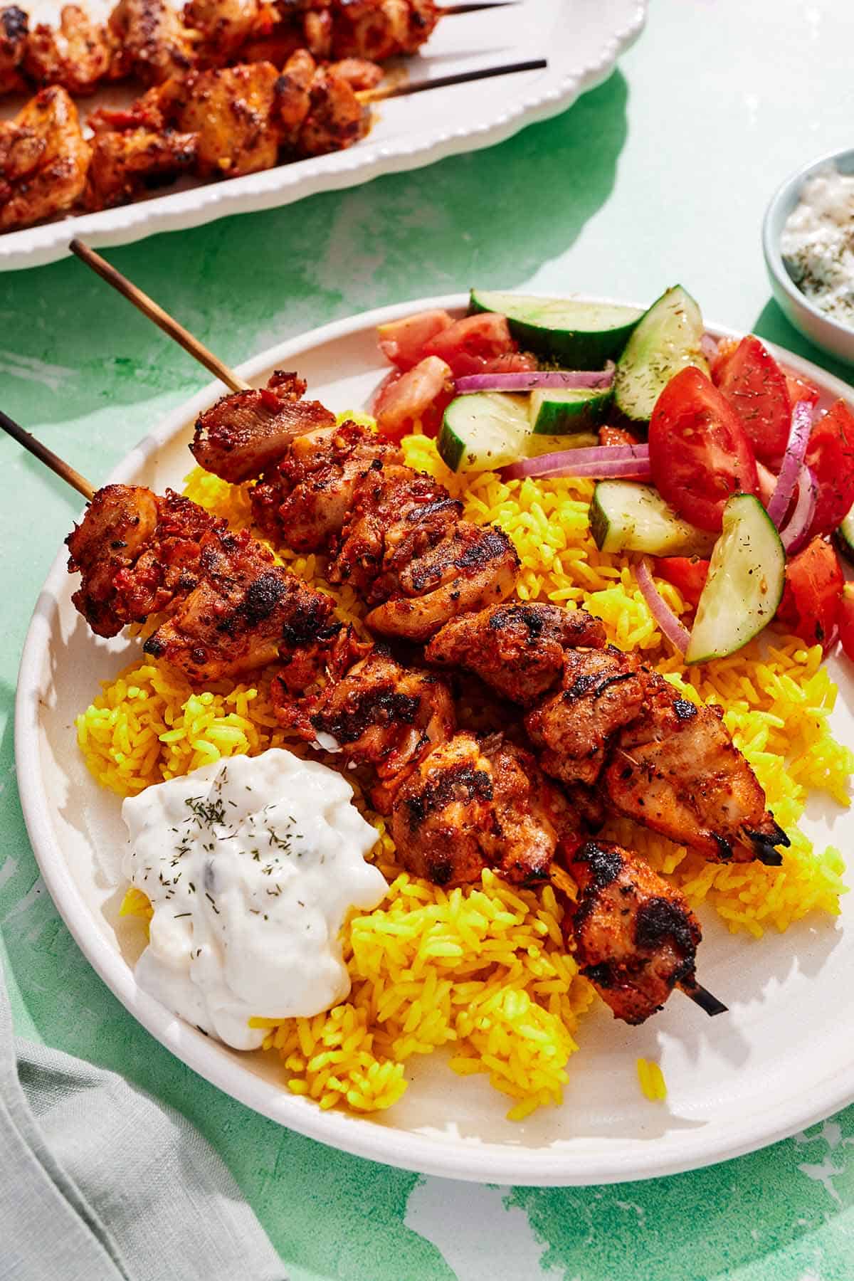 Turkish chicken skewers on a plate with rice, tzatziki and a cucumber and tomato salad.