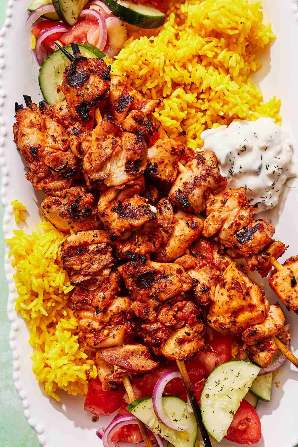 Turkish chicken skewers on a platter with rice, tzatziki and a cucumber and tomato salad.