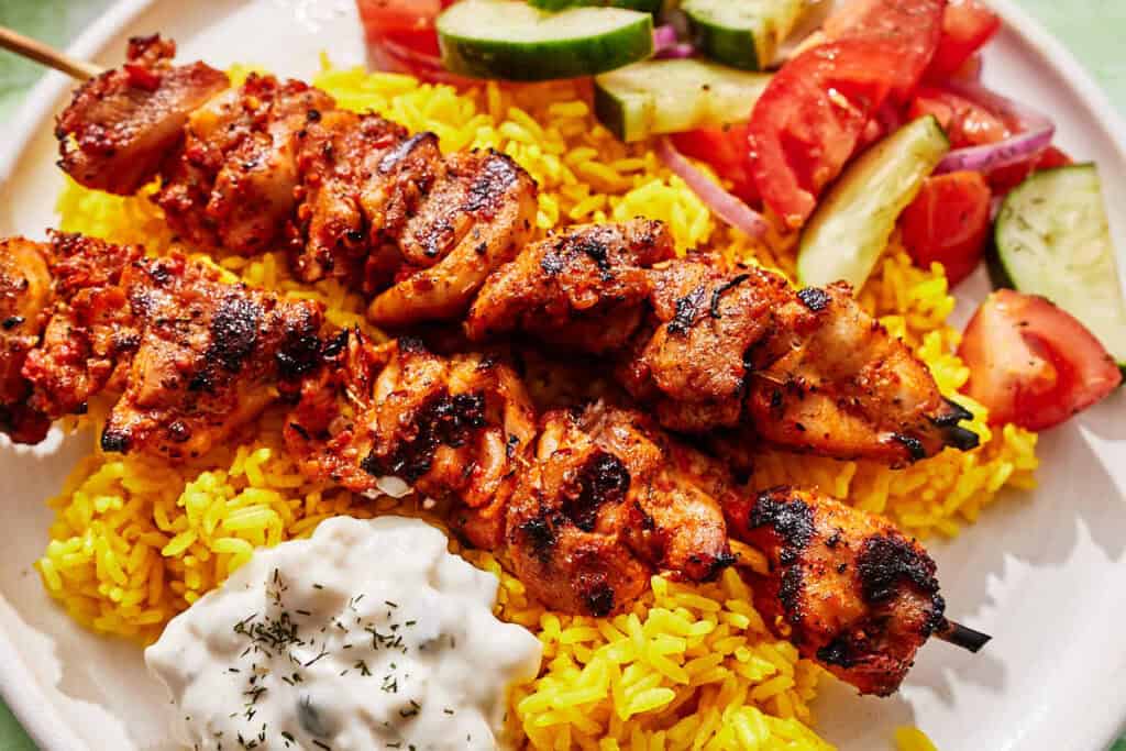 Turkish chicken skewers on a plate with rice, tzatziki and a cucumber and tomato salad.
