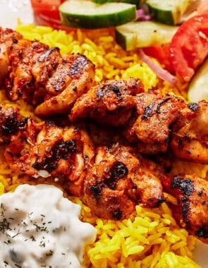 Turkish chicken skewers on a plate with rice, tzatziki and a cucumber and tomato salad.