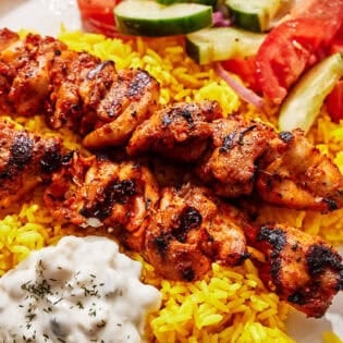 Turkish chicken skewers on a plate with rice, tzatziki and a cucumber and tomato salad.