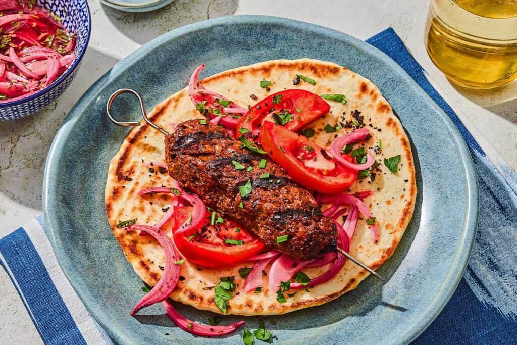 An adana kebab on a pita with pickled red onions and tomatoes. Next to this is a bowl of picked red onions and a glass of sparkling water.