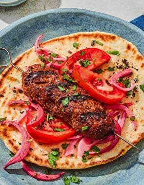 An adana kebab on a pita with pickled red onions and tomatoes. Next to this is a bowl of picked red onions and a glass of sparkling water.