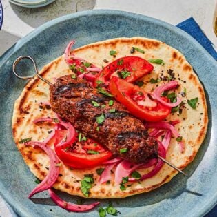 An adana kebab on a pita with pickled red onions and tomatoes. Next to this is a bowl of picked red onions and a glass of sparkling water.