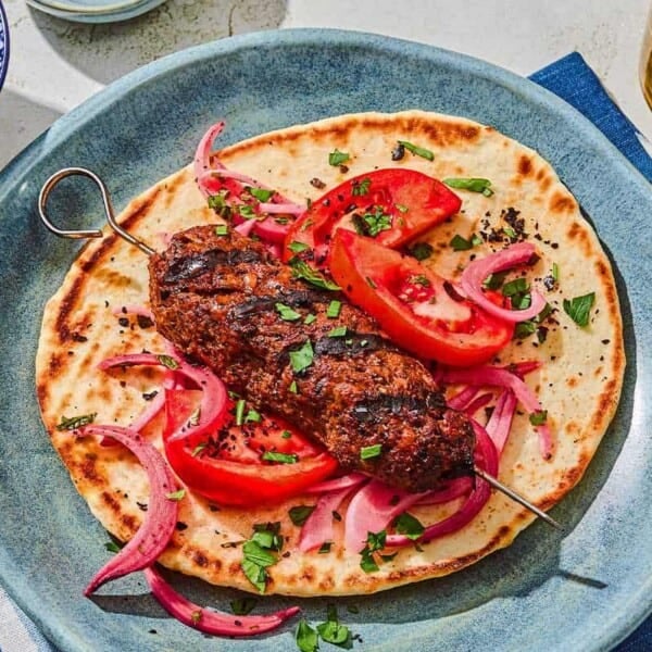 An adana kebab on a pita with pickled red onions and tomatoes. Next to this is a bowl of picked red onions and a glass of sparkling water.