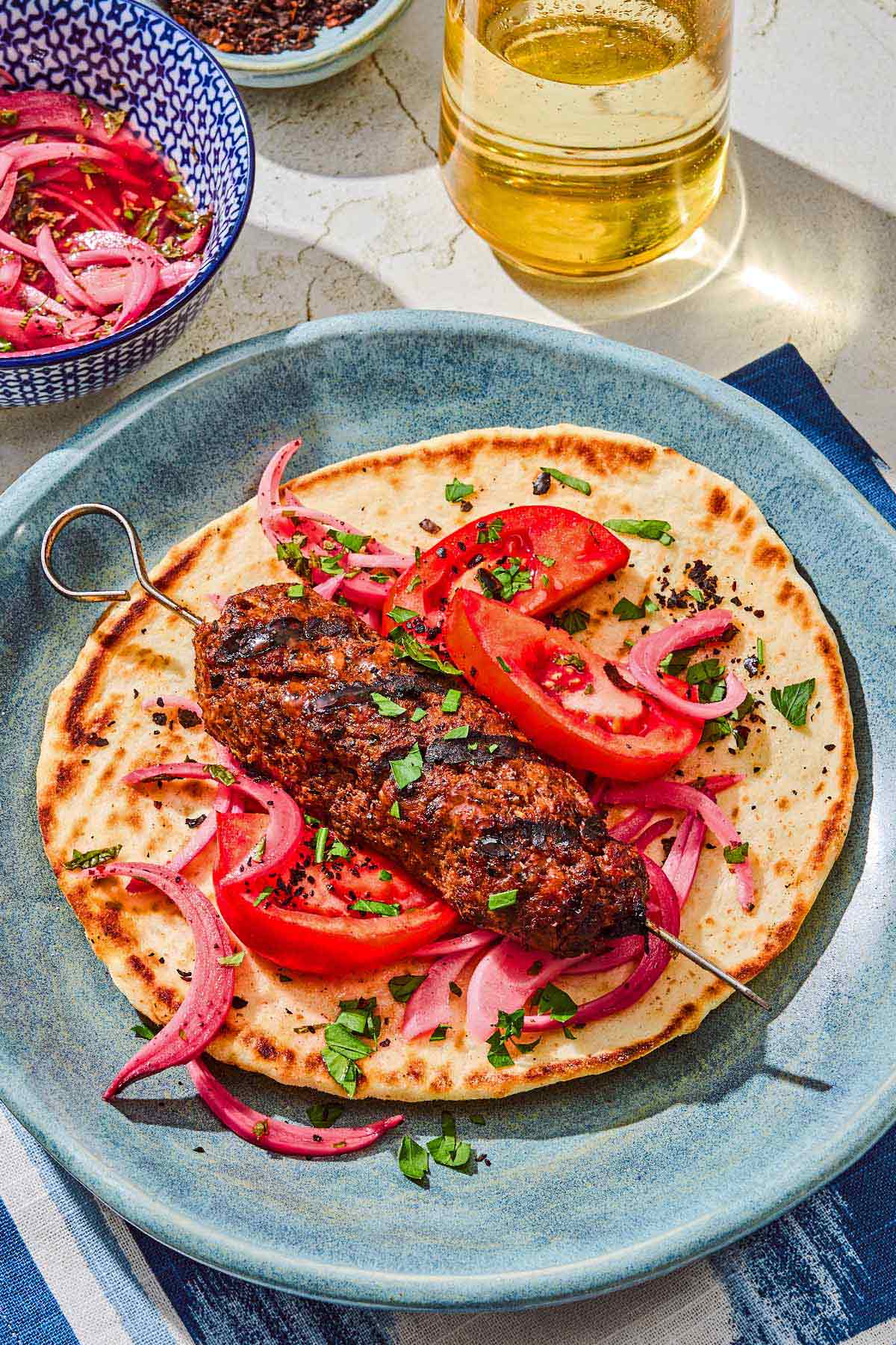 An adana kebab on a pita with pickled red onions and tomatoes. Next to this is a bowl of picked red onions and a glass of sparkling water.