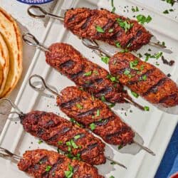 Several adana kebabs on a platter next to a stack of pita bread and bowls of pickled red onions, tomatoes and urfa biber.