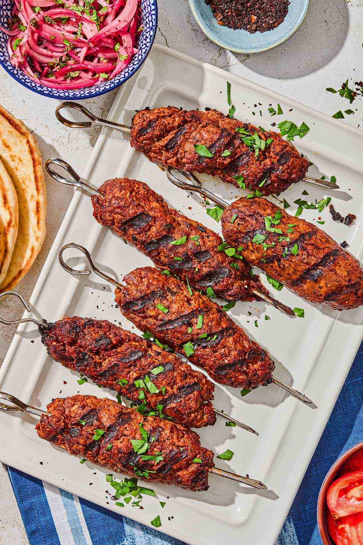 Several adana kebabs on a platter next to a stack of pita bread and bowls of pickled red onions, tomatoes and urfa biber.