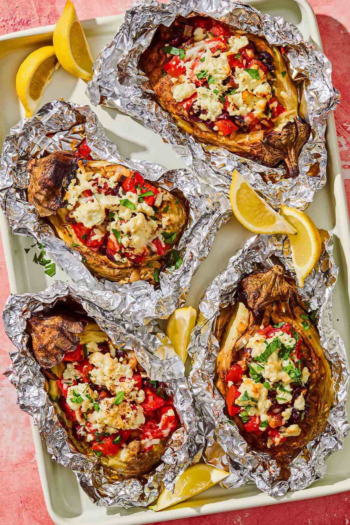 Four grilled eggplant parcels in foil on a platter with lemon wedges.