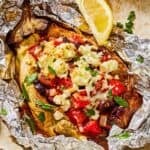 A grilled eggplant parcel in foil on a plate with a lemon wedge.