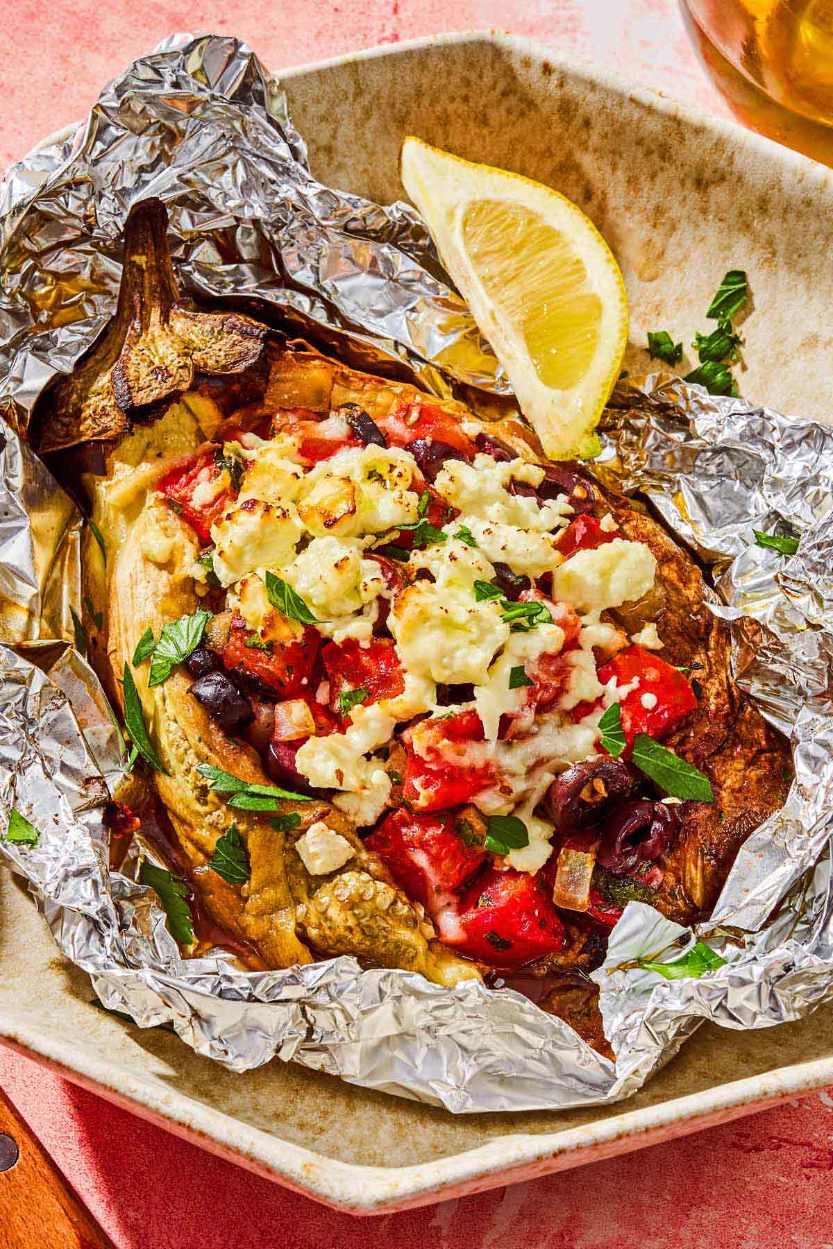 A grilled eggplant parcel in foil on a plate with a lemon wedge.