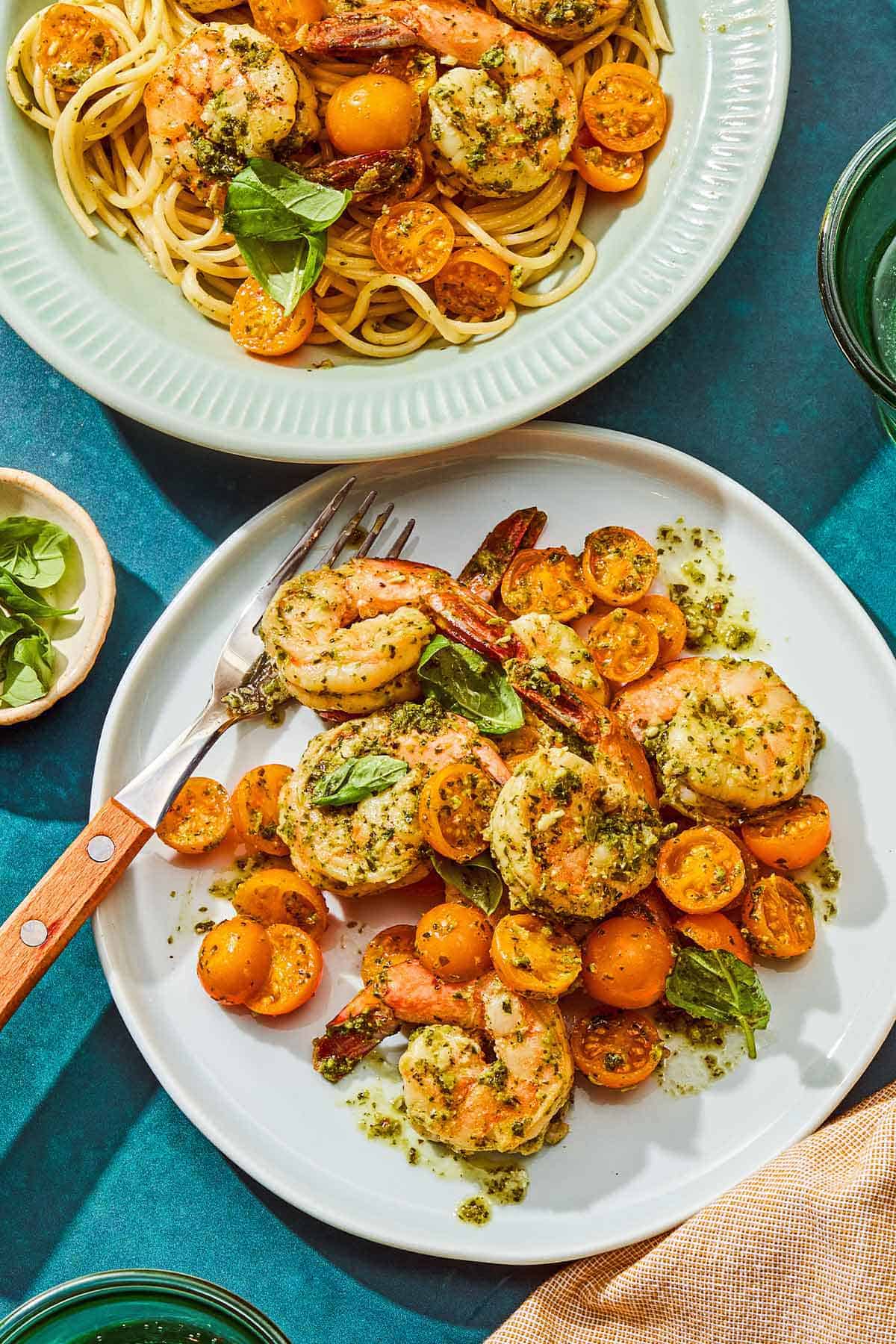A serving of pesto shrimp on a plate with a fork, next to a bowl with the rest of the shrimp.