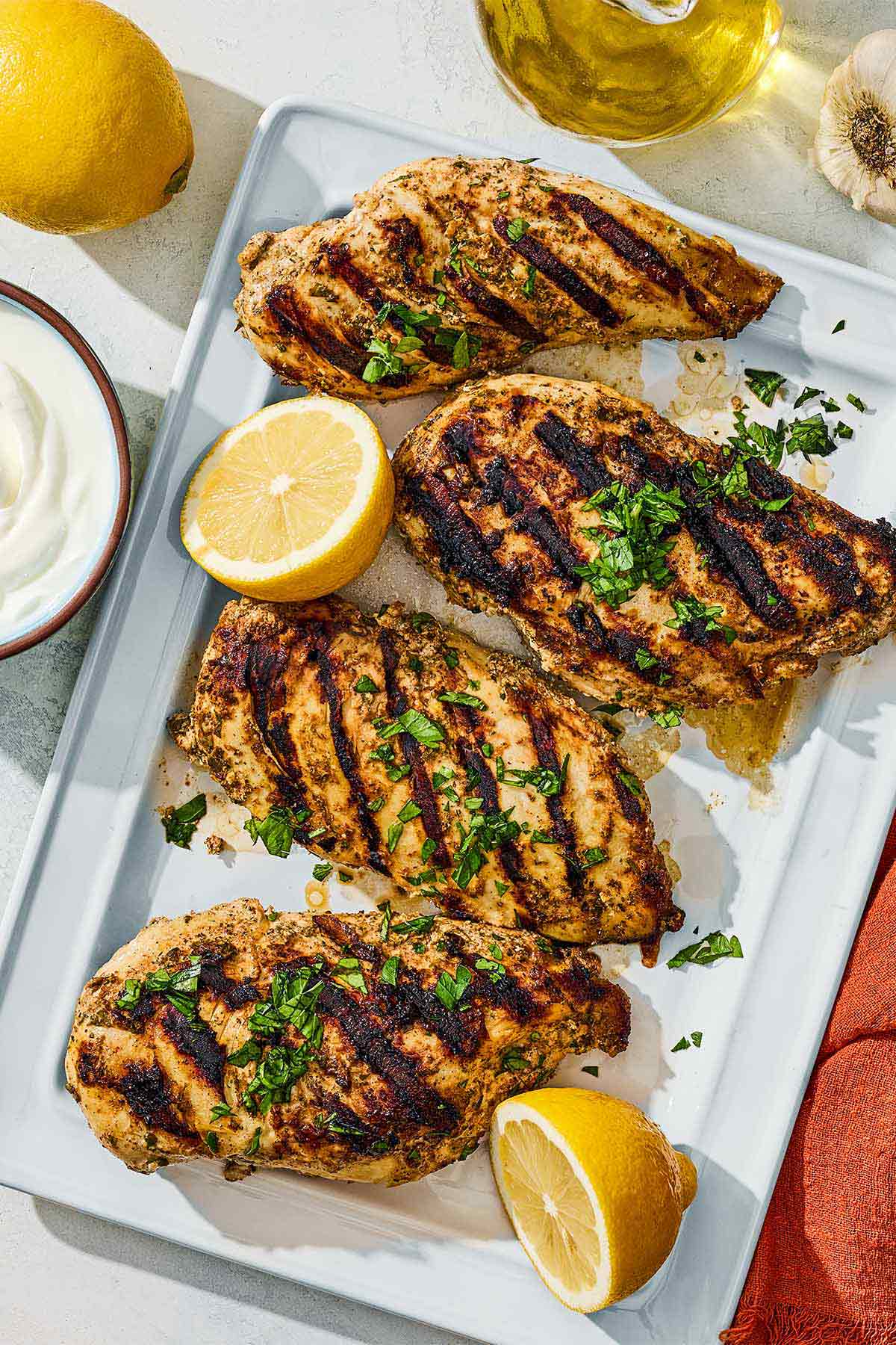 For grilled marinaded chicken breasts topped with parsley on a platter with lemon halves. Next to this is a bowl of yogurt, a lemon and a bottle of olive oil.