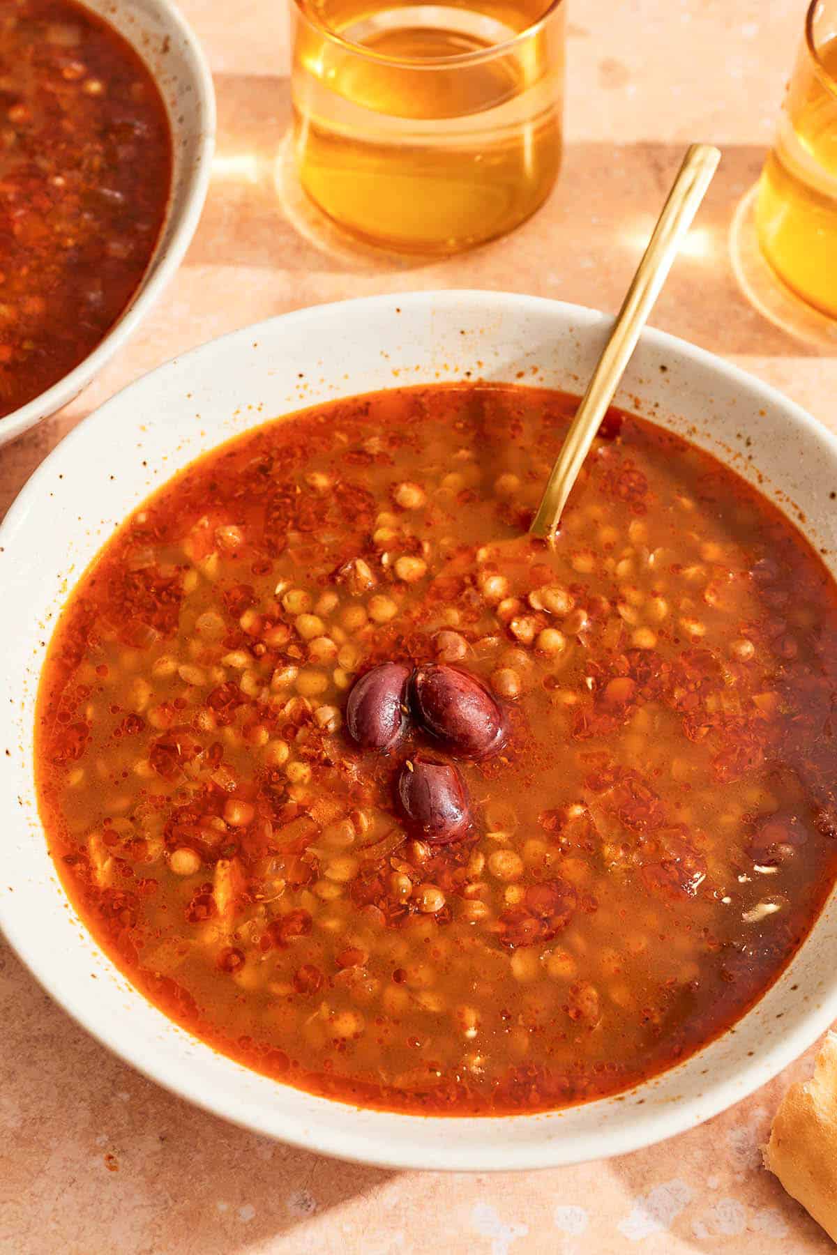 Greek lentil soup in a bowl with a spoon next to a few drinks and another bowl of the soup.