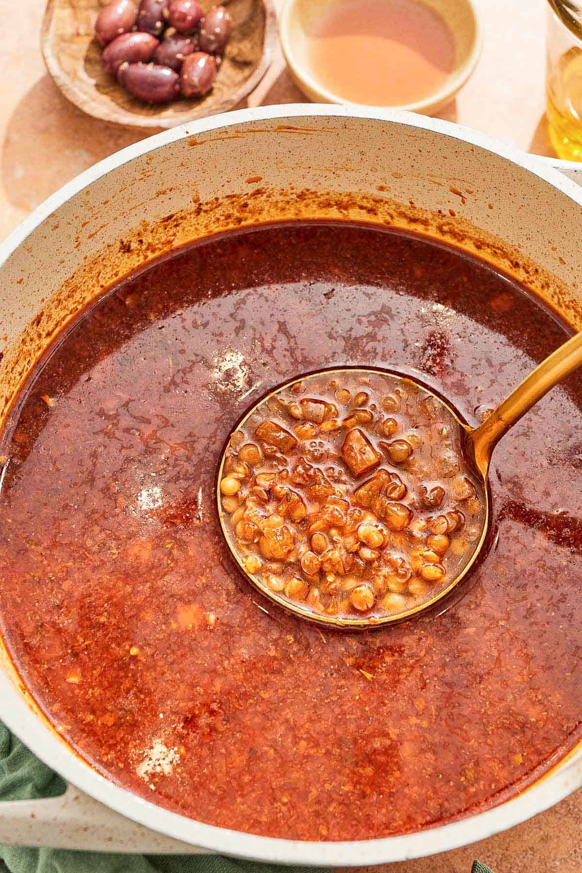 A ladleful of greek lentil soup being held over the rest of the pot of the soup.