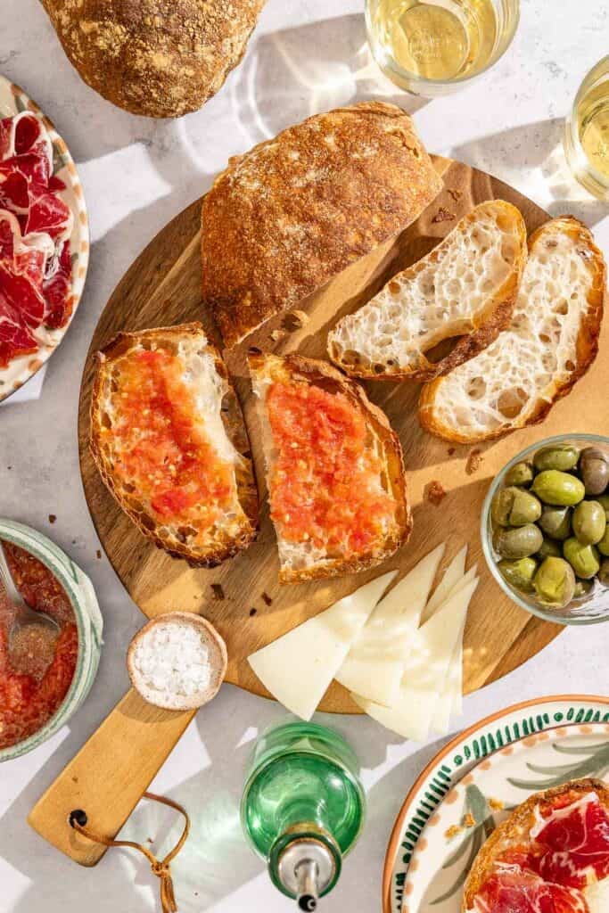 Slices of pan de cristal on a wooden board, some with a tomato topping. Surround this is salt, olives, cheese slices, olive oil, sliced meat and glasses of wine.