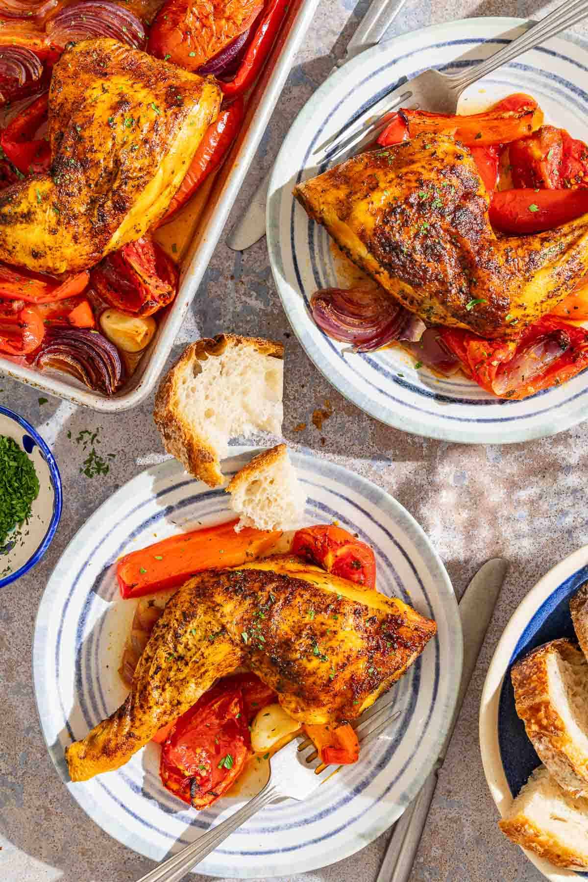 Two plates, each with a serving of Spanish paprika chicken and a fork. Next to these is the rest of the recipe on a sheet pan, a piece of crusty bread, and a bowl with more slices of bread.