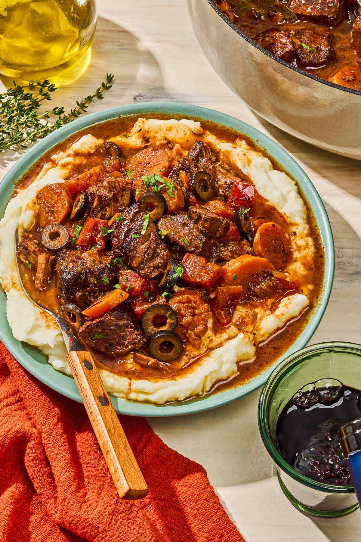 Daube provencal in a bowl with a fork next to a bottle of olive olive oil, a glass of red wine and the rest of the daube provencal in a pot.