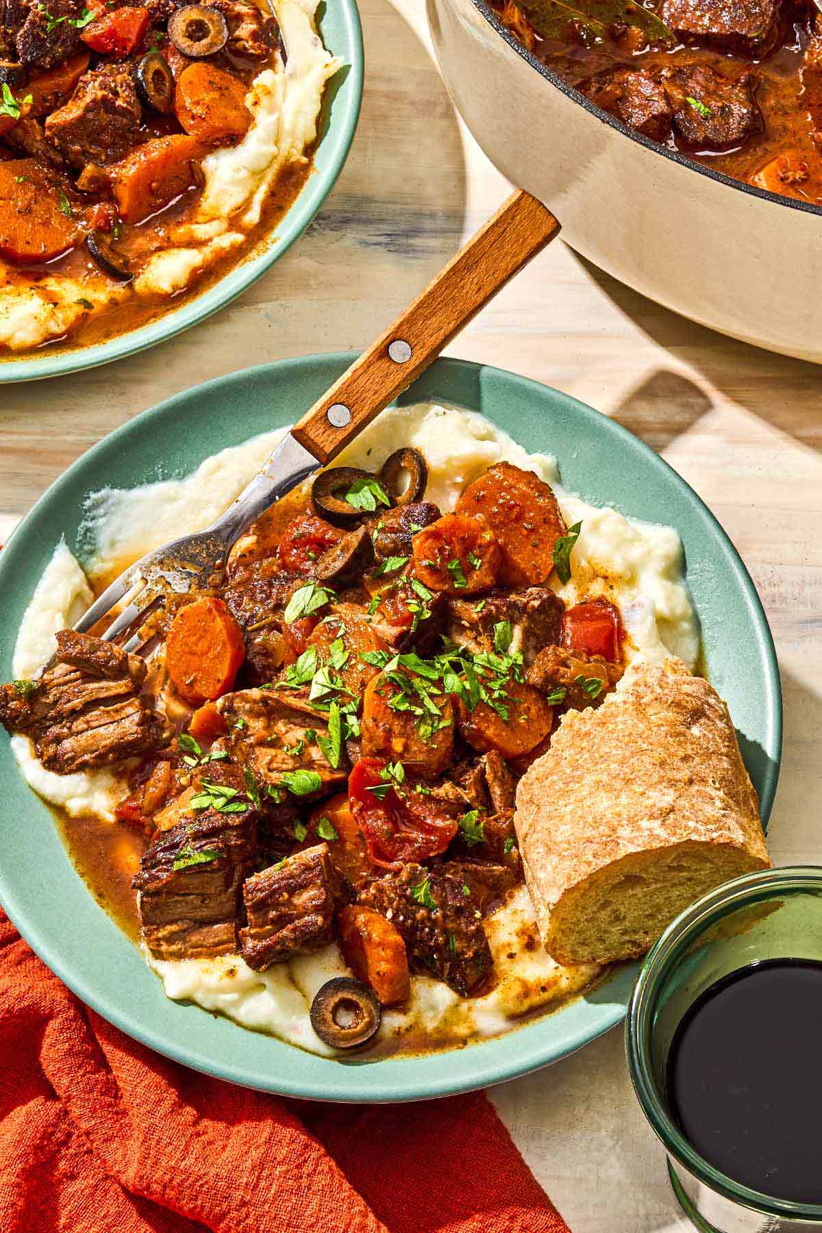 Daube provencal in a bowl with a fork and a slice of crusty bread next to another serving of the recipe in a bowl, the rest of the recipe in a pot, and a glass of red wine.
