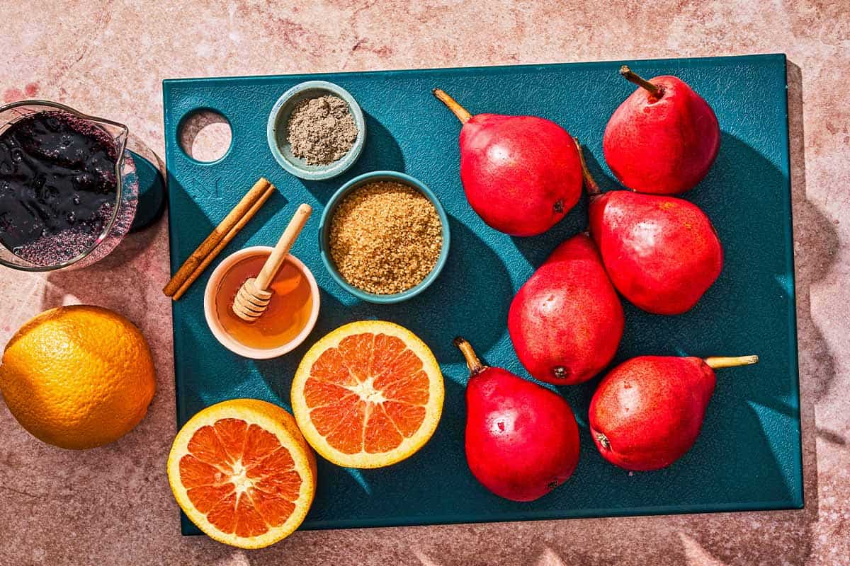 Ingredients for poached pears including pears, oranges, red wine, honey, sugar, a cinnamon stick, and ground cardamom.