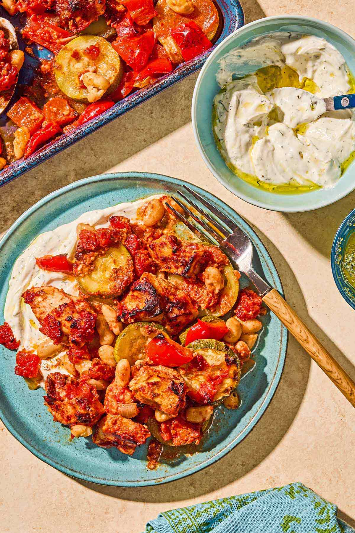 Sheet pan chicken and vegetables on a plate with a fork next to a bowl of Haydari with a spoon.