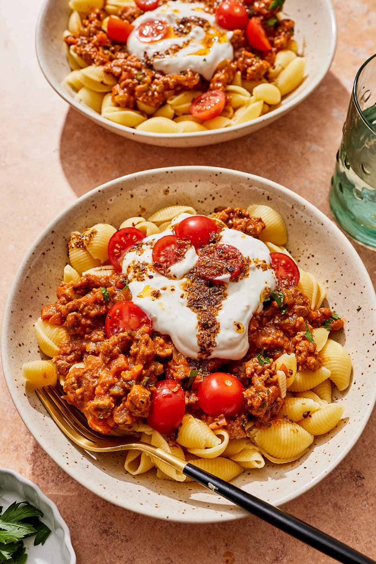Two bowls of Turkish pasta topped with a meat sauce, cherry tomatoes, yogurt sauce and spiced oil, one with a fork.