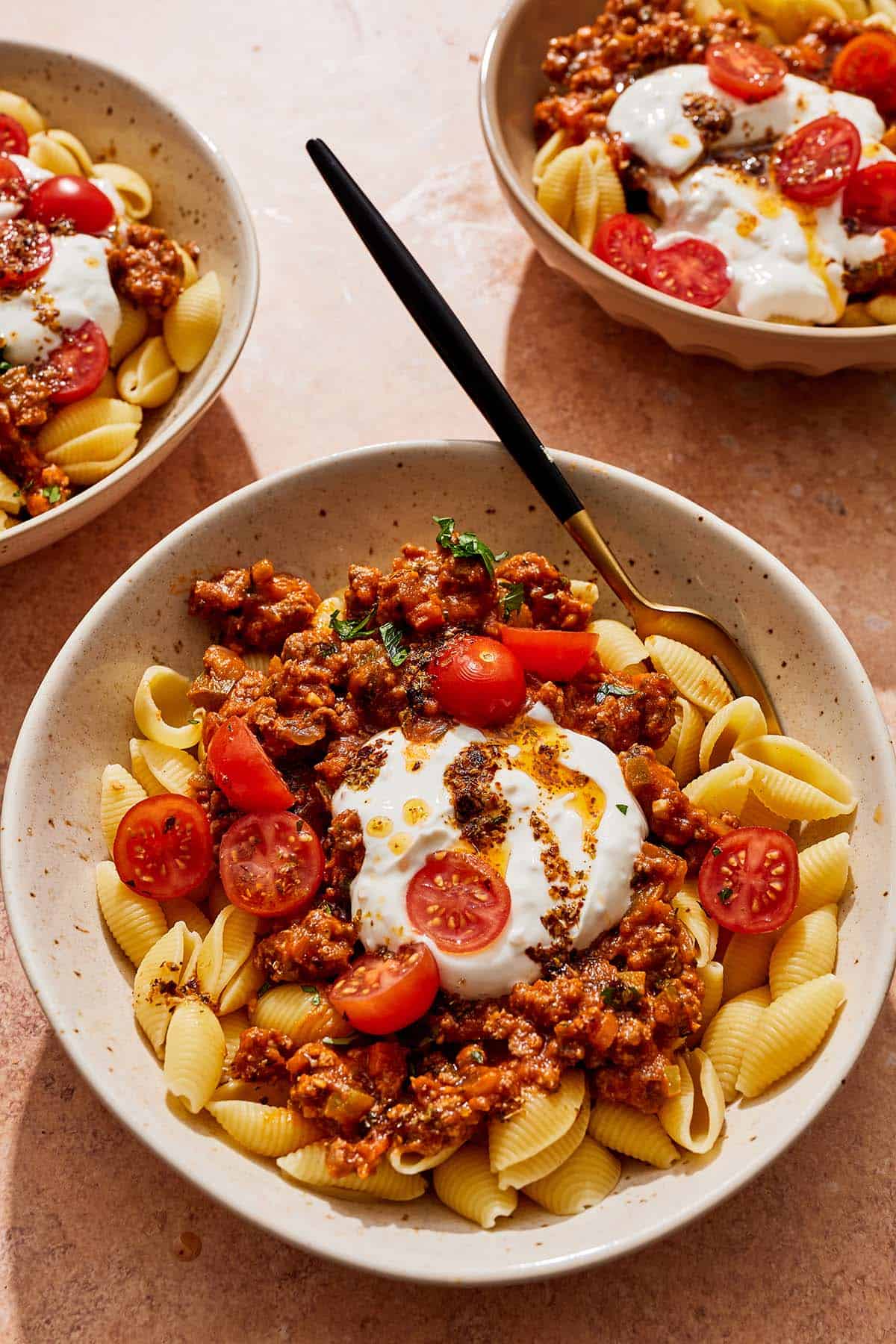 Three bowls of Turkish pasta topped with a meat sauce, cherry tomatoes, yogurt sauce and spiced oil in a bowl, one with a fork.